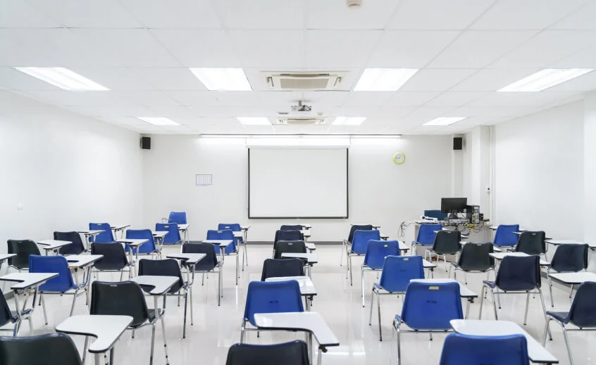 Empty classroom with blue and black chairs, whiteboard, projector screen, fluorescent lights, and ceiling AC, representing the education sector.