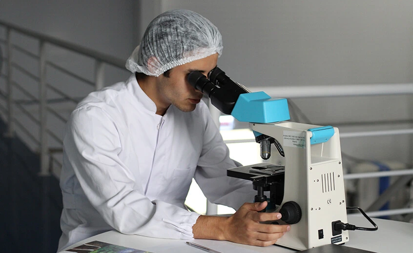 A laboratory researcher in a white lab coat and hairnet using a microscope. Illustrates the healthcare sector.